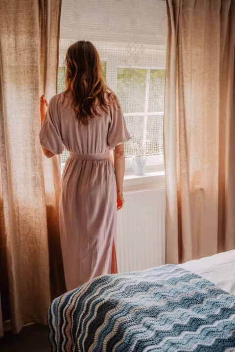 Osoba patrzy przez okno zasłonięte zasłonami.